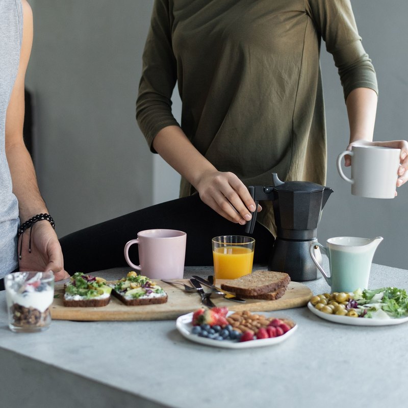 Breakfast of avocado toasts, berries, yoghurt and orange juice on a concrete counter while a woman holds a mug