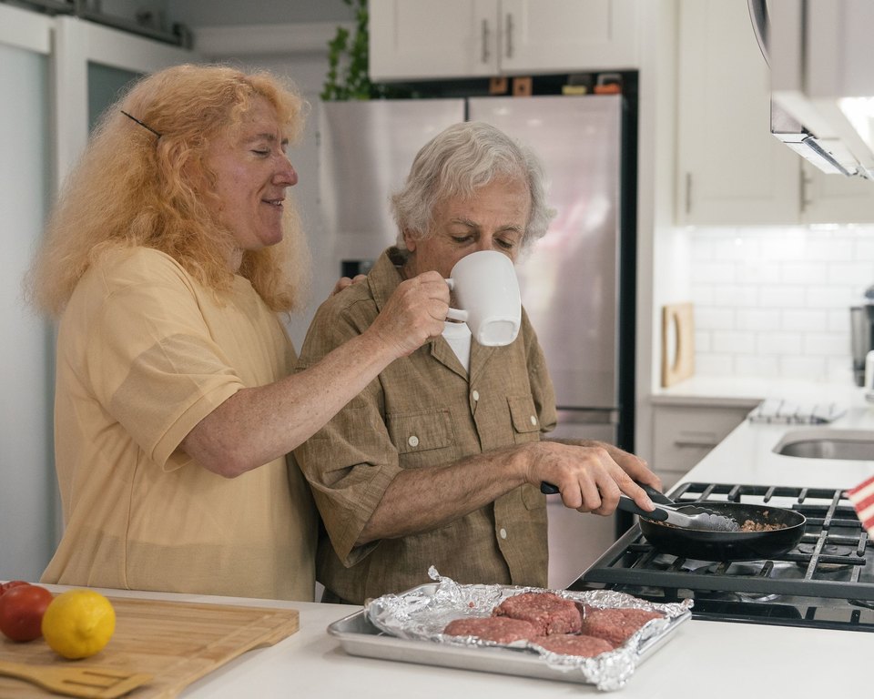Woman standing behind a man as he drinks from a mug and cooks at a gas stove