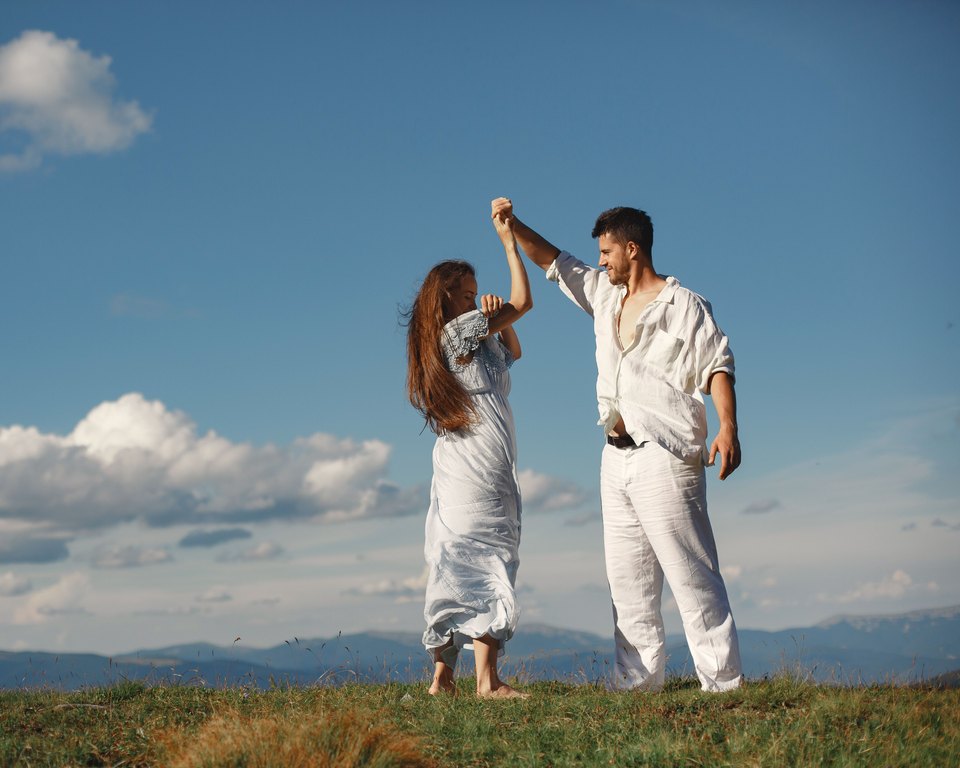 Man in white spinning a woman in a blue dress by the hand on a grassy hilltop under a blue sky