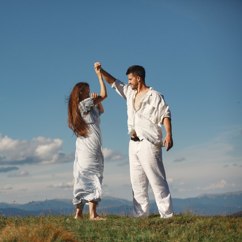 Man in white spinning a woman in a blue dress by the hand on a grassy hilltop under a blue sky