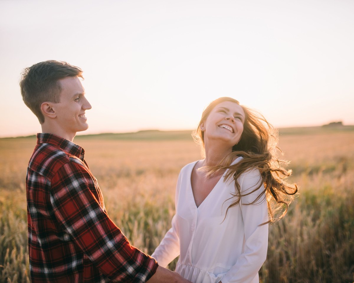 Smiling man holding hands with a laughing woman whose hair is blowing in the wind, in a golden wheat field at sunset