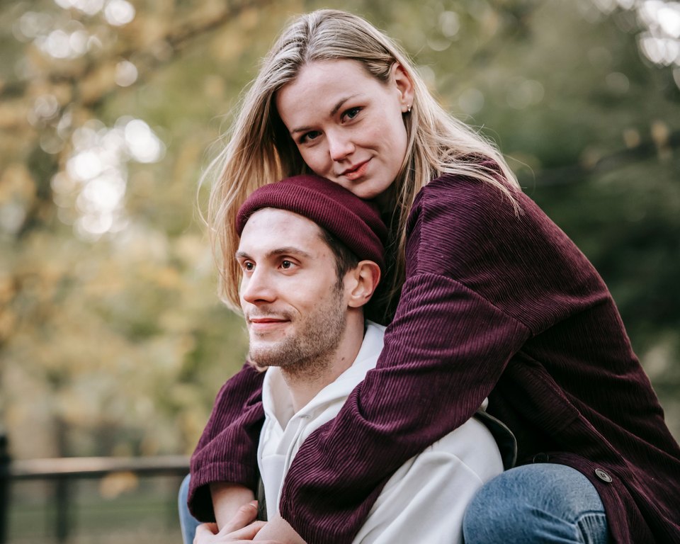 Woman in a burgundy jacket hugging a smiling man from behind on a piggyback ride in an autumn park