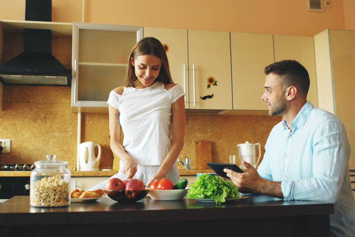 Woman slicing vegetables in a kitchen while a man sits at the counter with a tablet