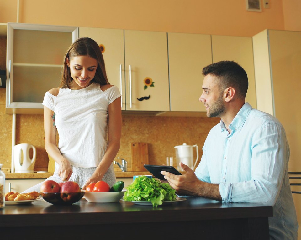 Woman slicing vegetables in a kitchen while a man sits at the counter with a tablet