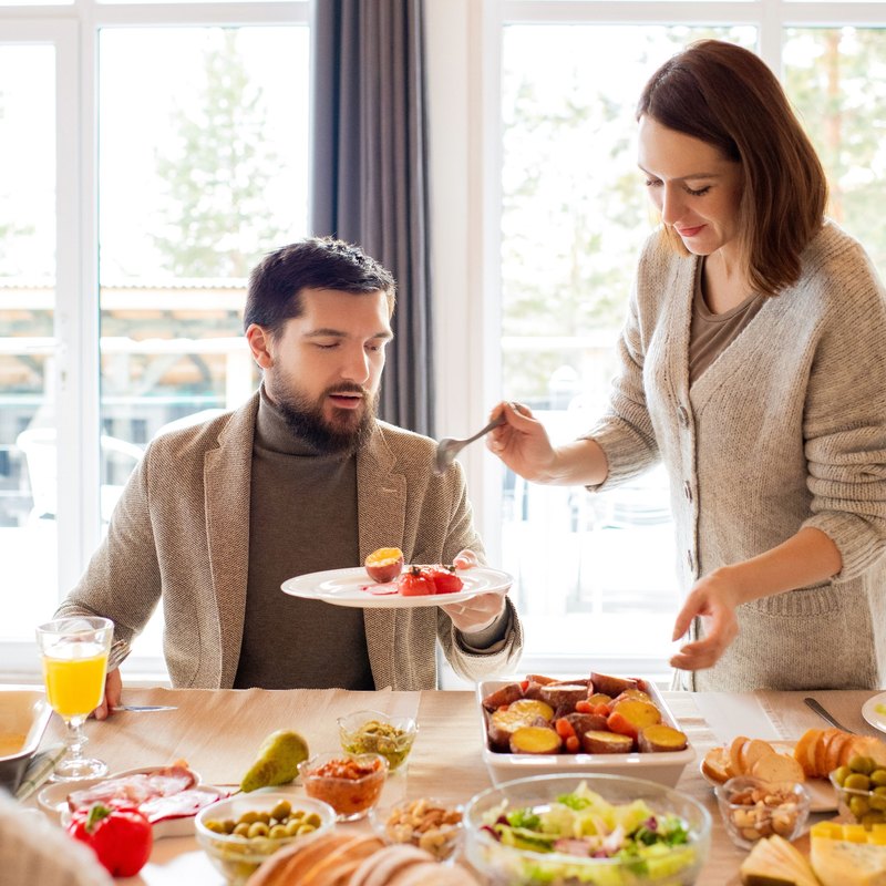 Woman serving food with a fork to a seated man at a table spread with salad, vegetables and bread