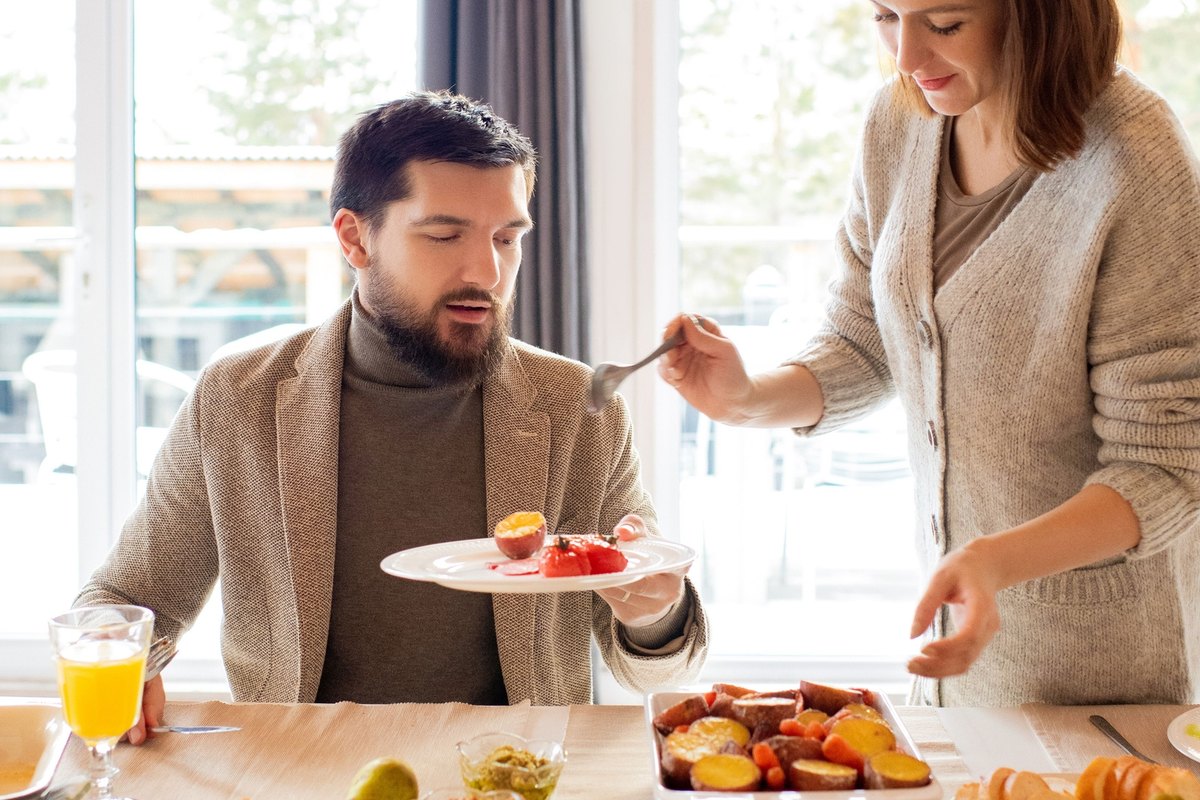 Woman serving food with a fork to a seated man at a table of salad, vegetables and bread