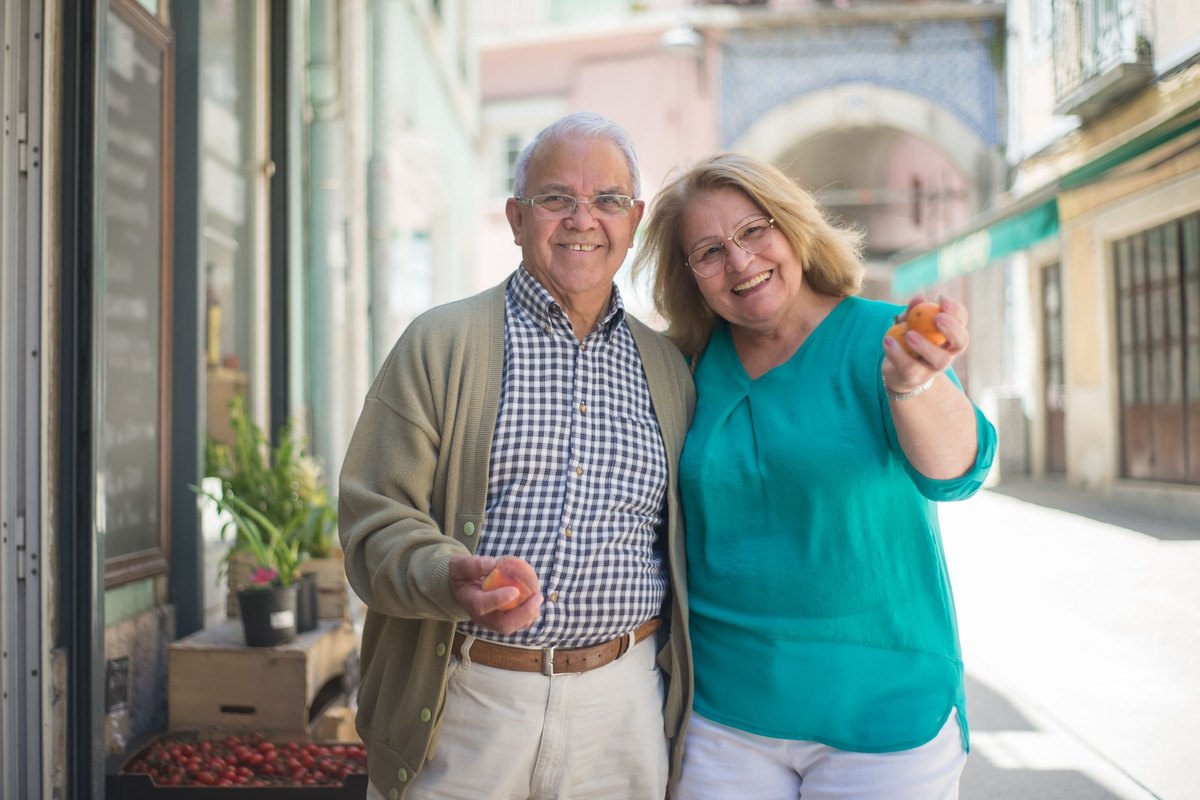 Smiling older couple holding fresh peaches and apricots outside a grocery on a sunny street