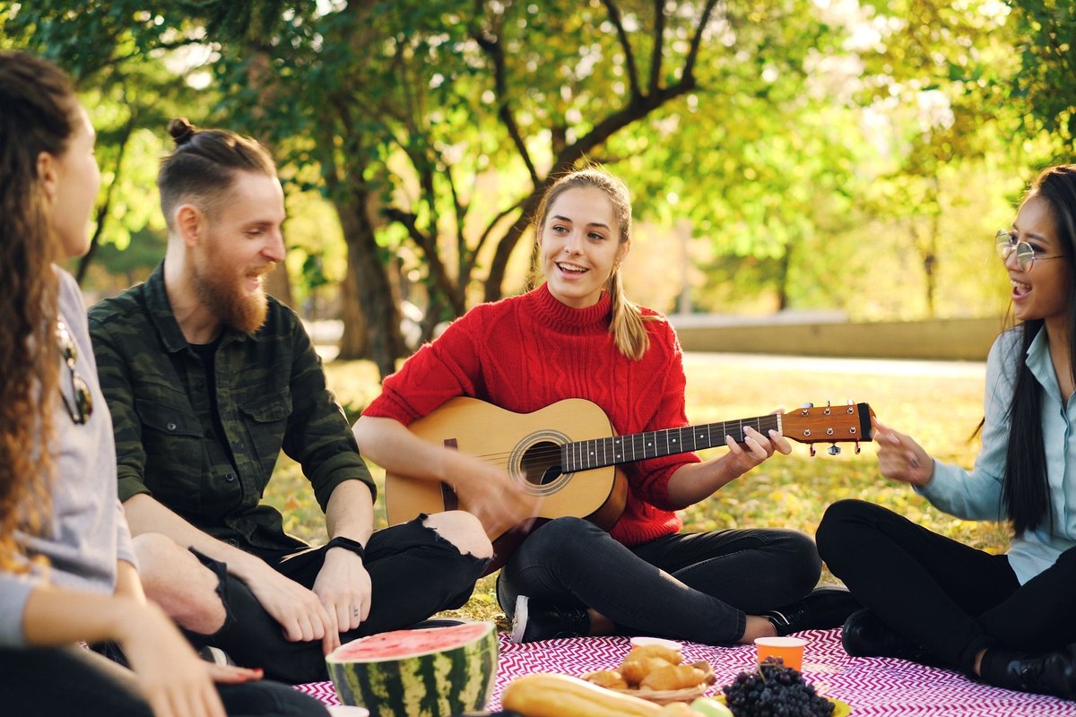 Four friends singing along to a guitar on a picnic blanket in a park