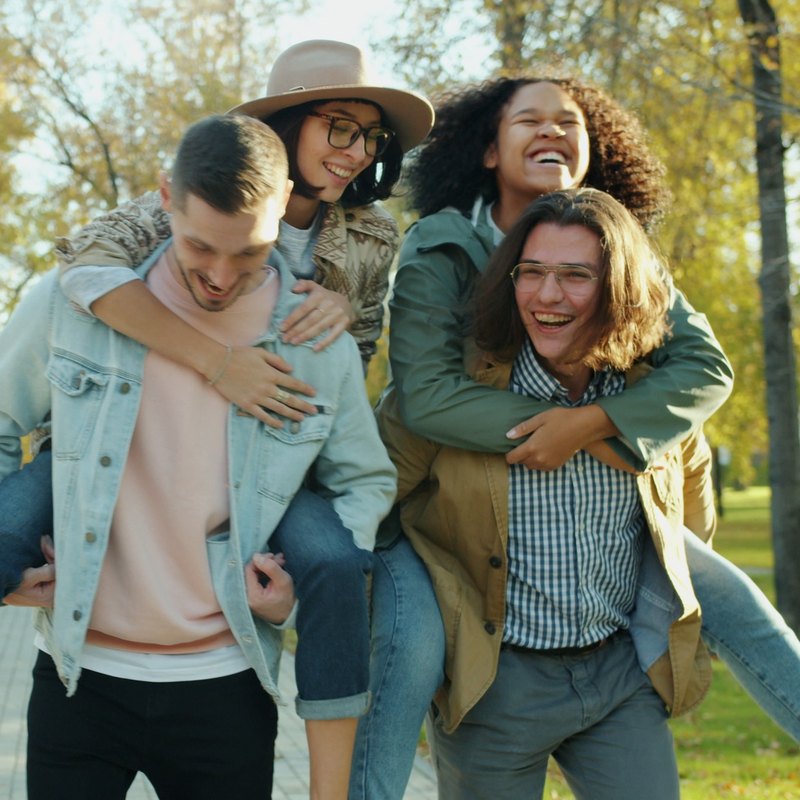 Laughing friends giving each other piggyback rides along a park path in autumn