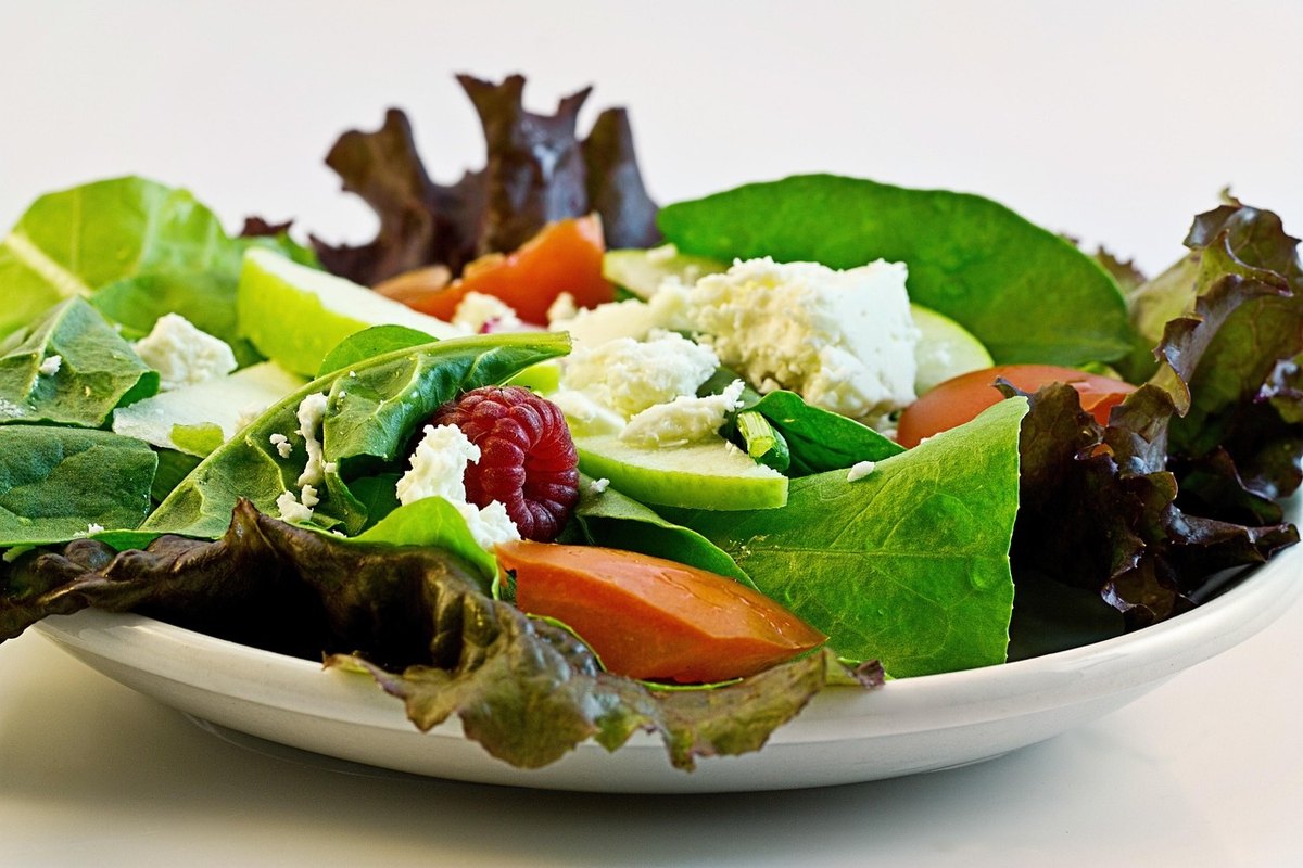 White plate of spinach and red-leaf lettuce with feta, apple, tomato and a raspberry