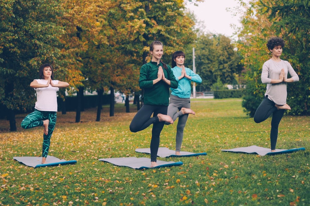 Four women balancing in tree pose with palms together on yoga mats in an autumn park