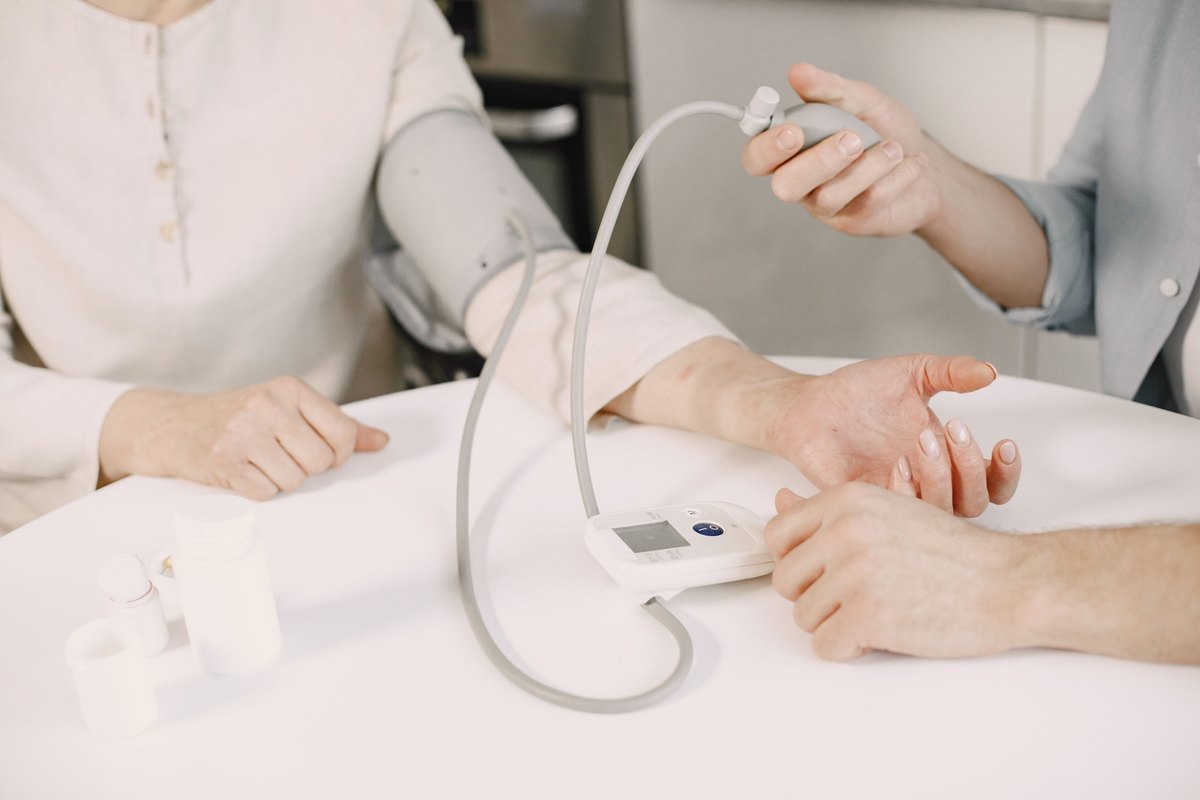 Person squeezing the bulb of a blood pressure cuff on a seated woman's arm at a white table
