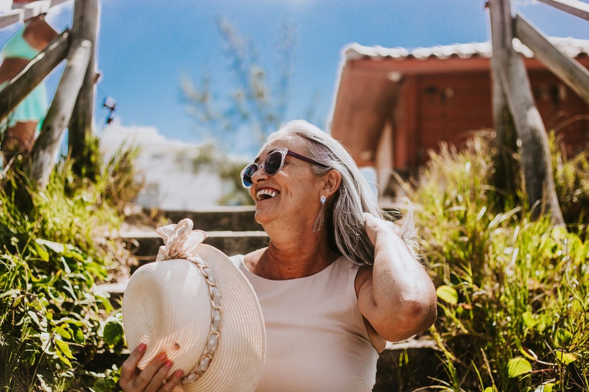 Laughing older woman in sunglasses holding a straw hat while sitting on steps among tall grass