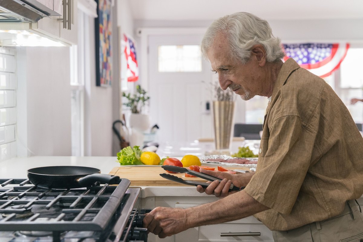 Man holding tongs at a gas stove in a bright kitchen