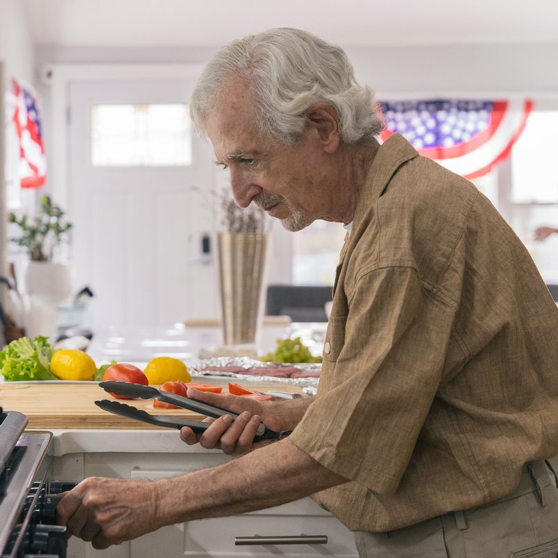 Man holding tongs at a gas stove in a bright kitchen