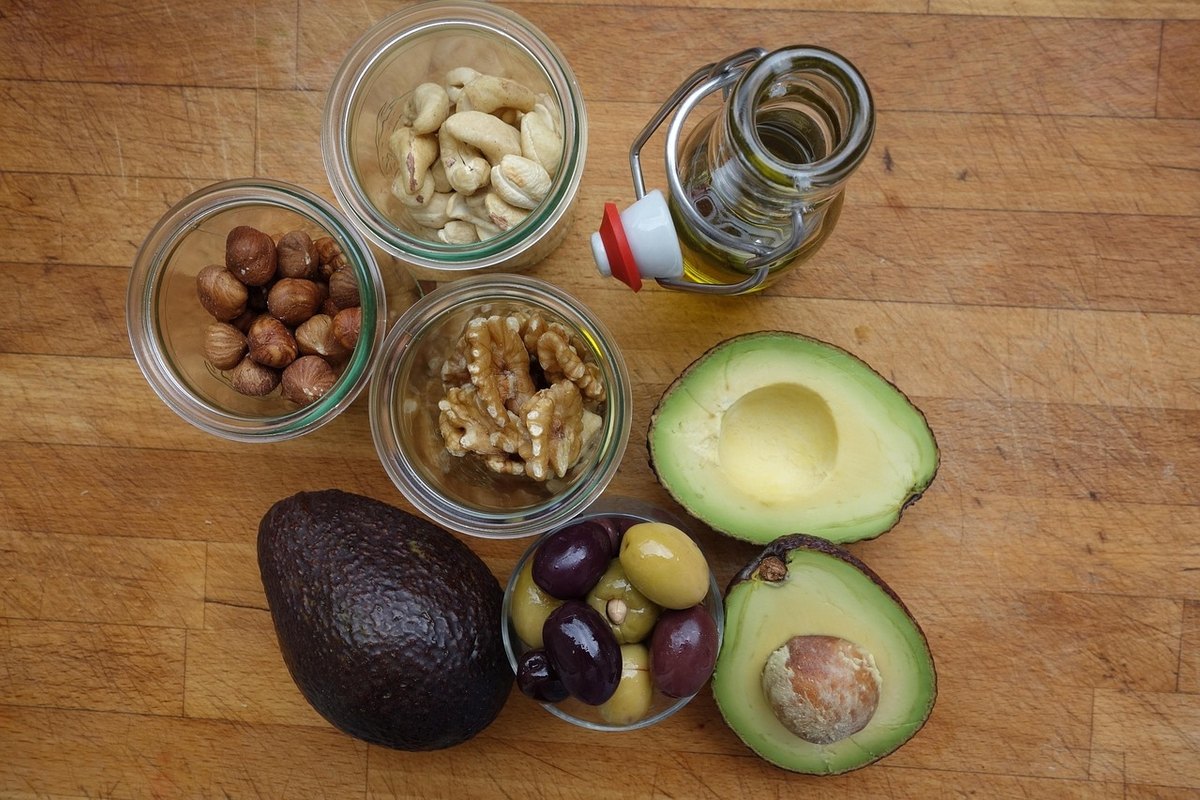 Jars of cashews, hazelnuts and walnuts beside olive oil, olives and avocado halves on a wooden board