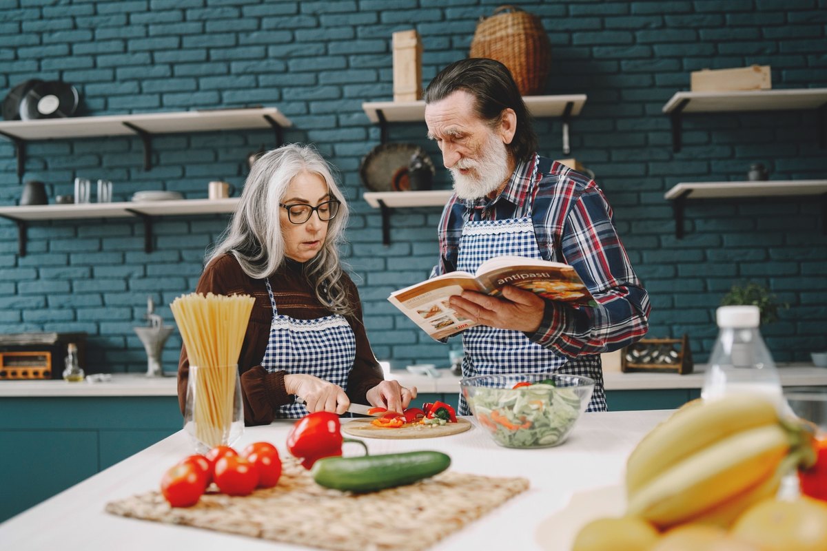Older couple in matching aprons preparing a salad and reading a cookbook in a kitchen