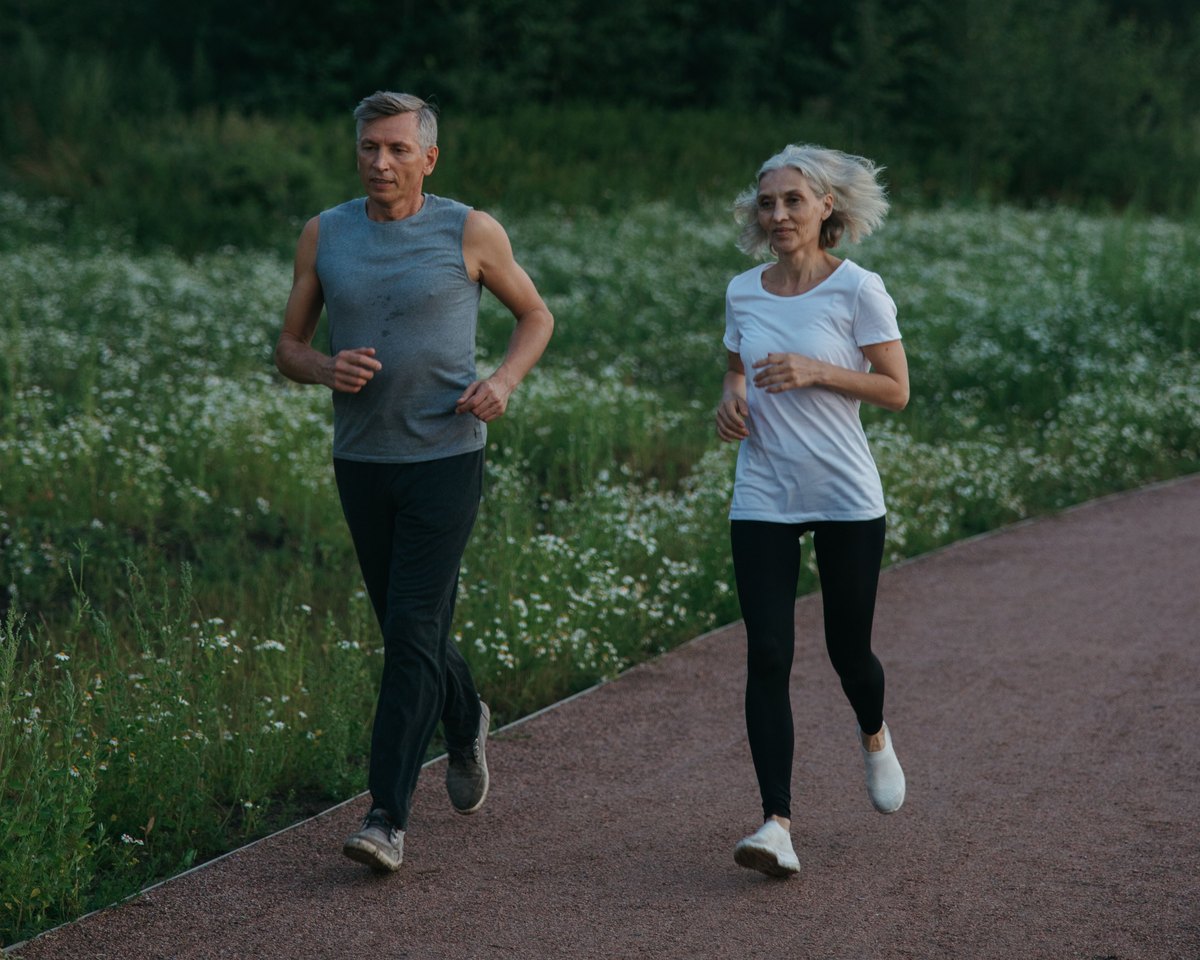 Older man and woman jogging side by side on a gravel path beside a meadow of white flowers
