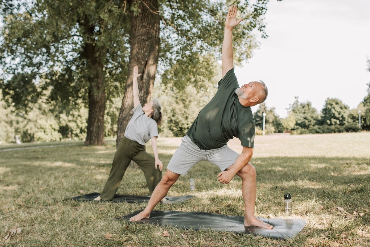 Older man and woman doing the same side-stretch pose on exercise mats in a park