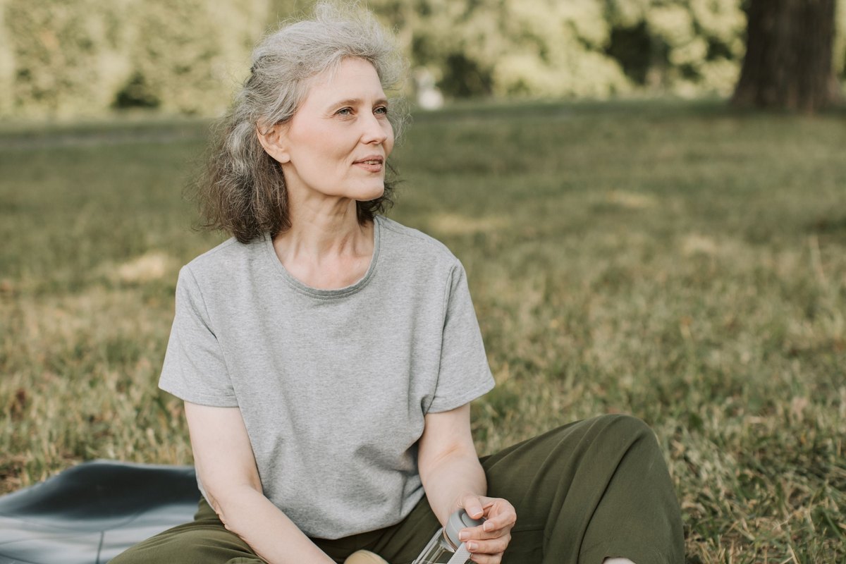 Older woman with gray hair sitting on a yoga mat in a park, holding a water bottle