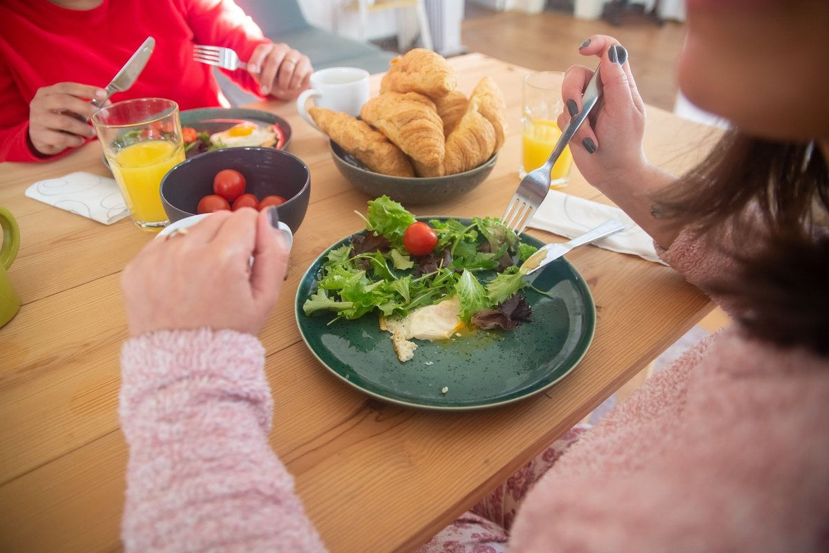 Two people eating a breakfast of mixed salad and a fried egg at a wooden table with croissants and orange juice