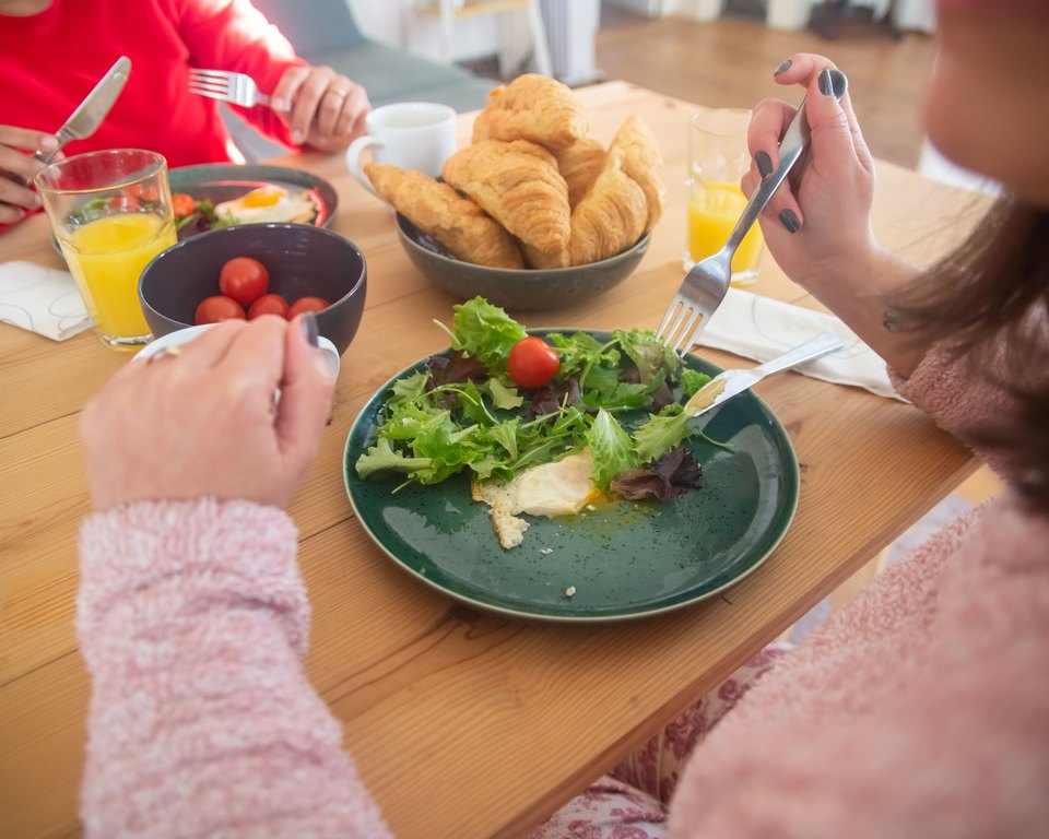 Two people eating a breakfast of mixed salad and a fried egg at a wooden table with croissants and orange juice