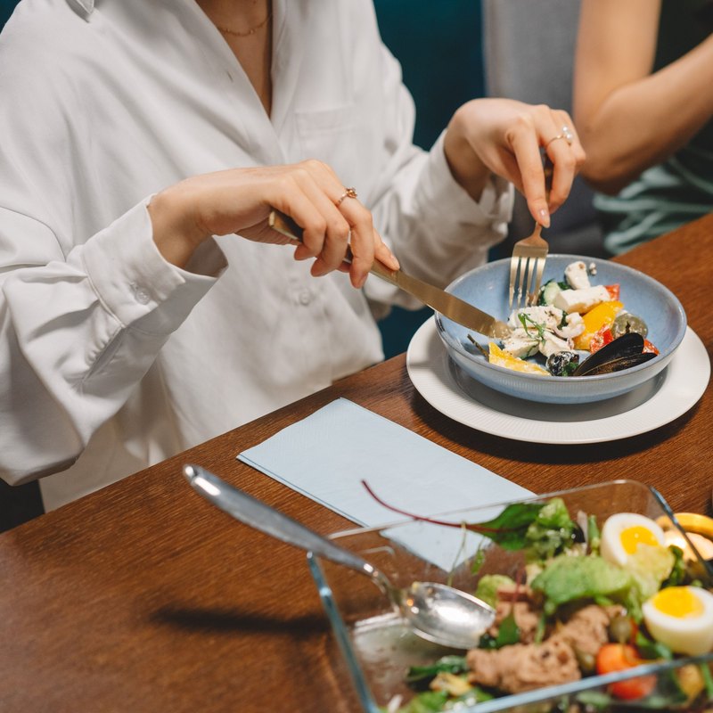 Woman in a white shirt eating a salad from a blue bowl at a table laid with several dishes