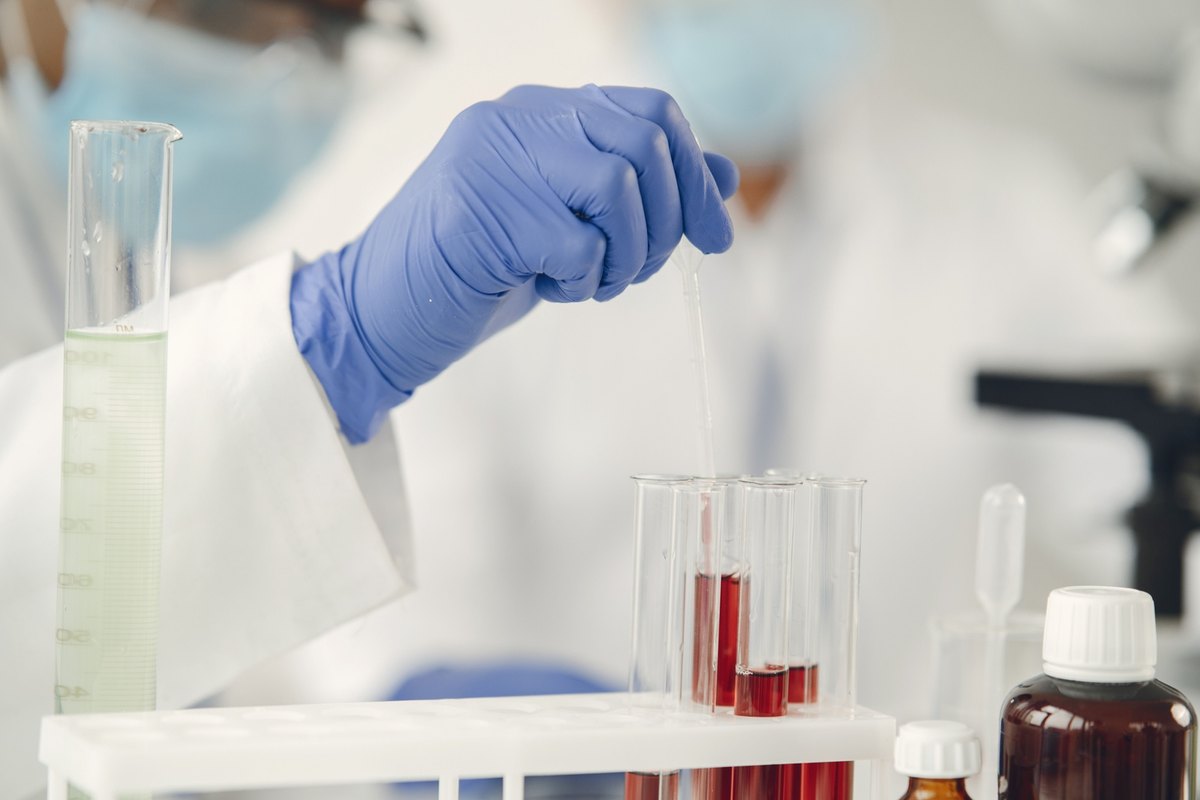 Gloved hand holding a pipette over a rack of test tubes with red liquid in a laboratory