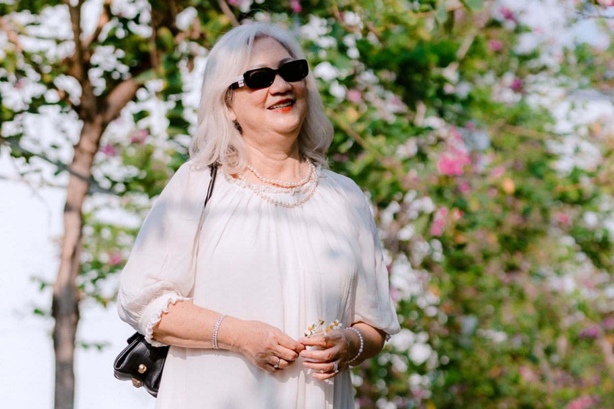Smiling older woman in a white dress and sunglasses holding a sprig of flowers under flowering trees