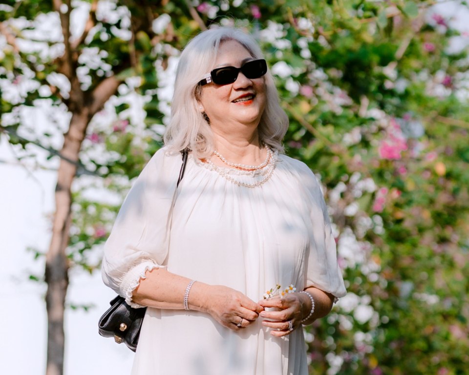 Smiling older woman in a white dress and sunglasses holding a sprig of flowers under flowering trees