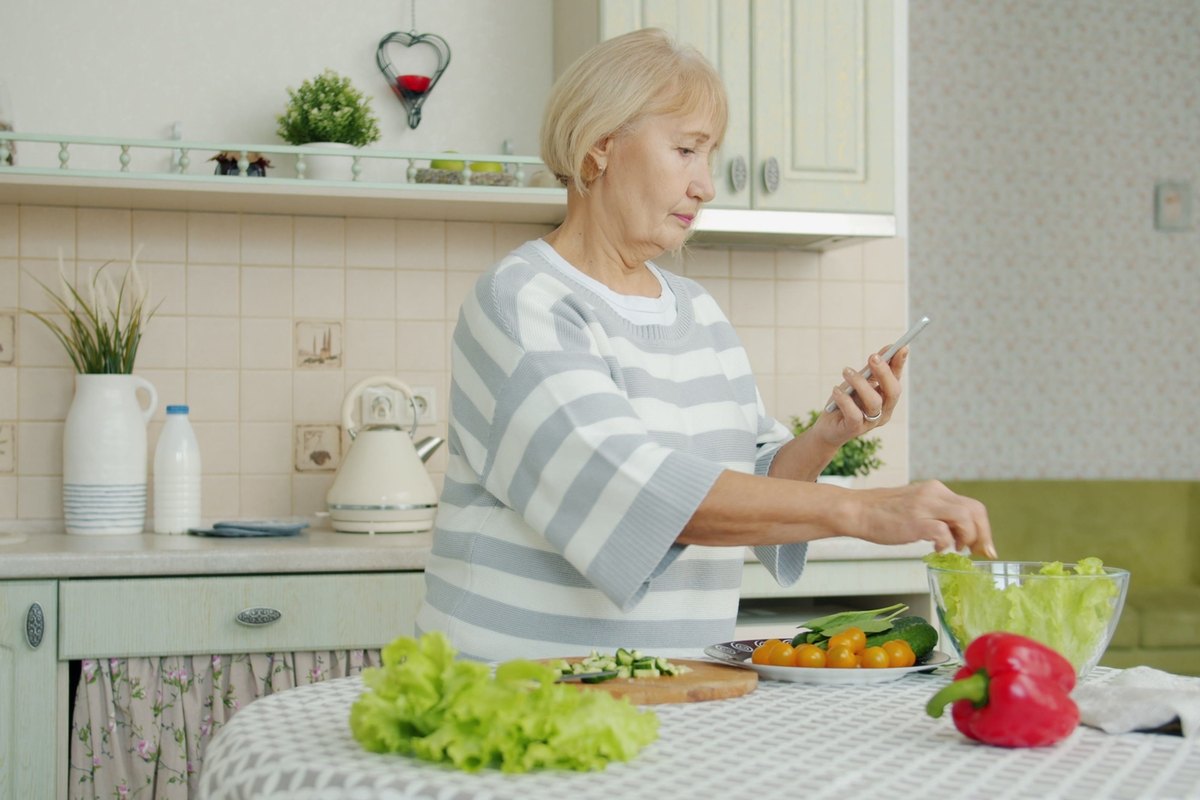 Older woman in a striped sweater making a salad in a kitchen while holding a phone