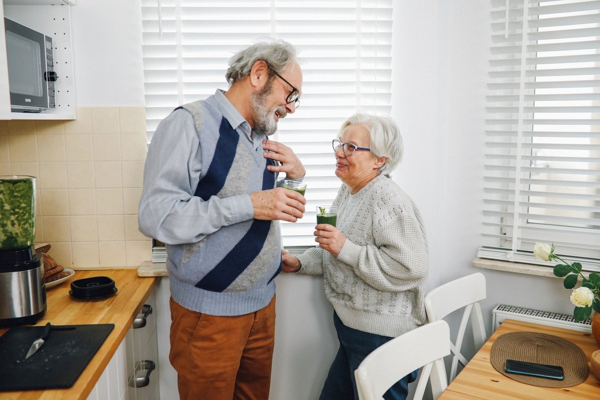 Older man with a gray beard and an older white-haired woman holding glasses of green smoothie by a window