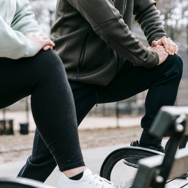 Two people in lunge stretches with their hands on their knees beside a park bench