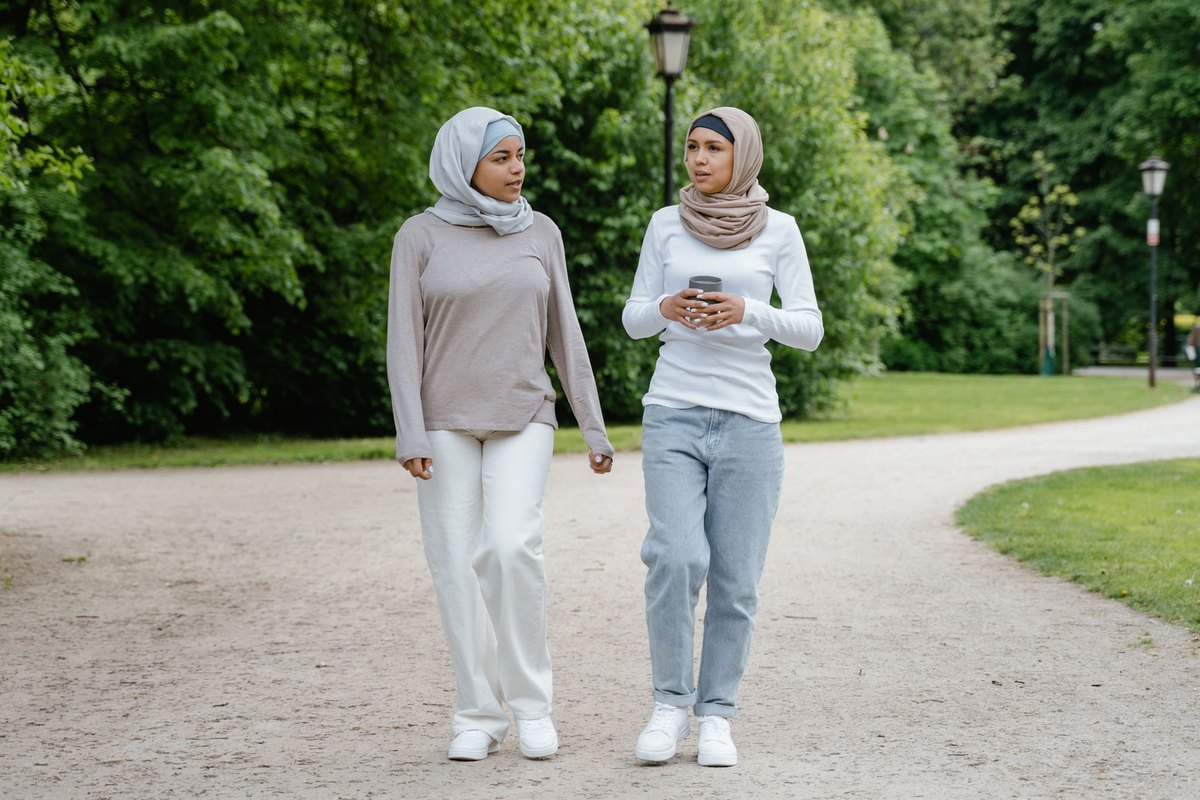 Two women in light head scarves talking as they walk along a gravel path in a leafy park