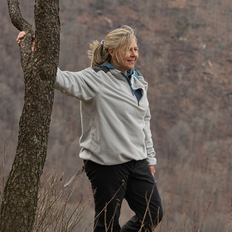 Smiling woman with windblown hair resting a hand on a pine tree at a mountain overlook