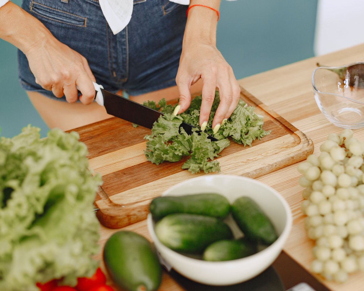 Woman in denim shorts slicing lettuce on a wooden cutting board beside bowls of cucumbers, grapes and other fresh produce