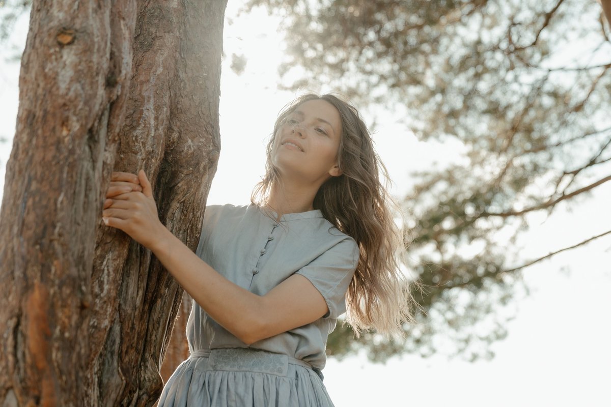 Woman in a pale blue blouse holding the trunk of a pine tree and looking up with a soft smile