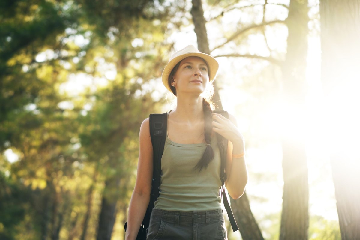 Woman in a straw hat and tank top with a backpack looking into the distance in a sunlit pine forest