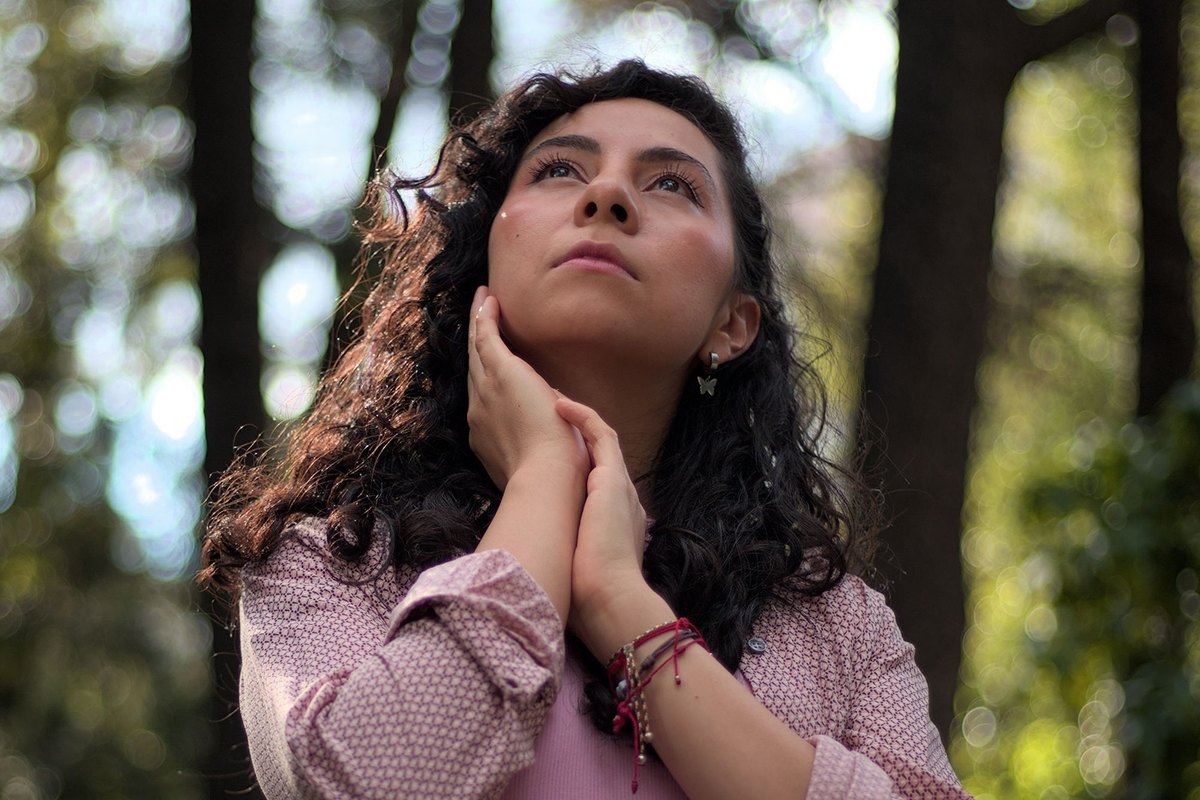 Woman with long curly hair looking up in a forest, one hand resting on her jaw