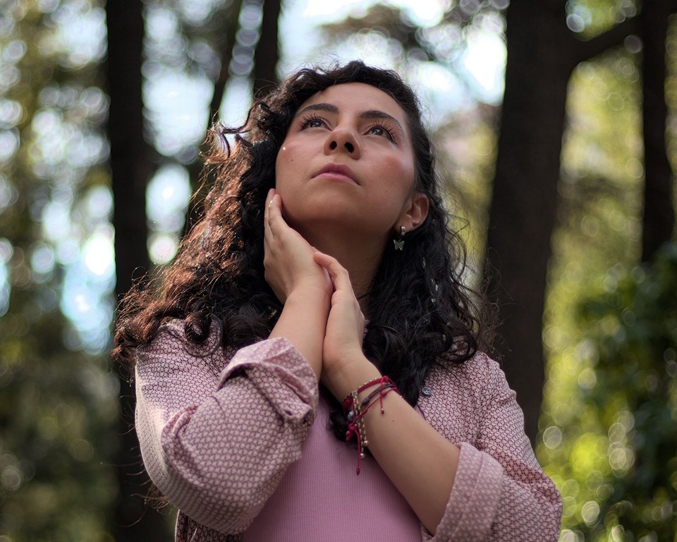 Woman with long curly hair looking up in a forest, one hand resting on her jaw
