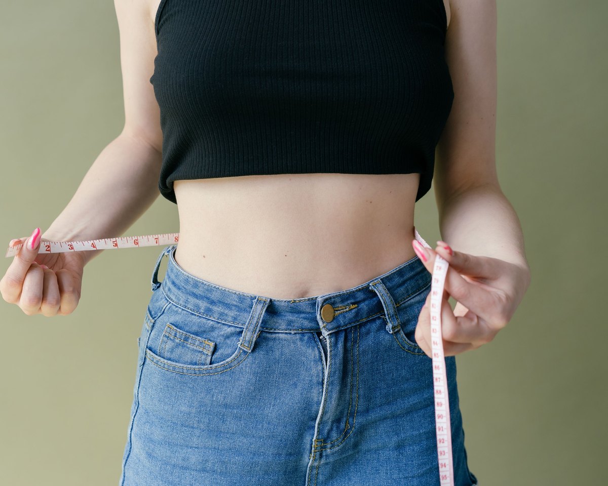 Woman in a black crop top and jeans holding a measuring tape against her waist