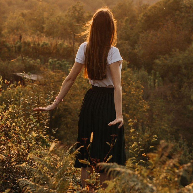 Woman seen from behind with her arms out, standing among plants on a hillside at sunset
