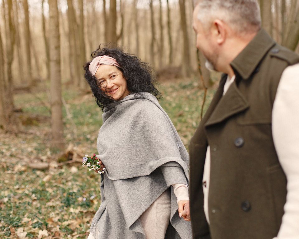 Smiling woman in a gray shawl holding flowers and looking back at a man as they walk through a spring forest