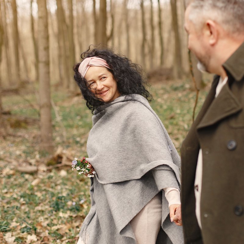 Smiling woman in a gray shawl holding flowers and looking back at a man as they walk through a spring forest