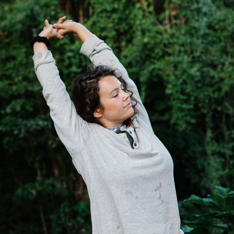 Woman with closed eyes stretching both arms overhead in front of green leaves in a park