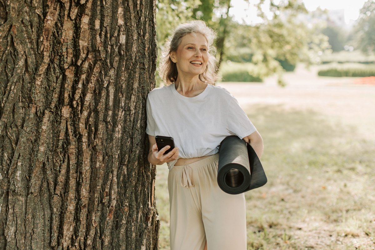 Smiling gray-haired woman leaning on a tree trunk in a park, holding a phone and a rolled yoga mat