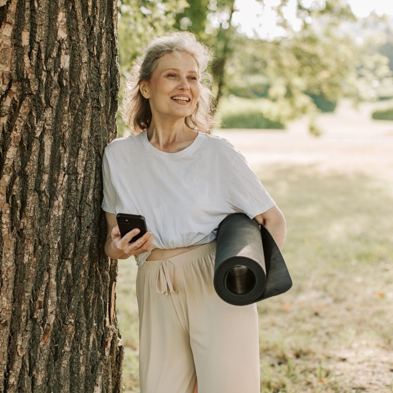 Smiling gray-haired woman leaning on a tree trunk in a park, holding a phone and a rolled yoga mat
