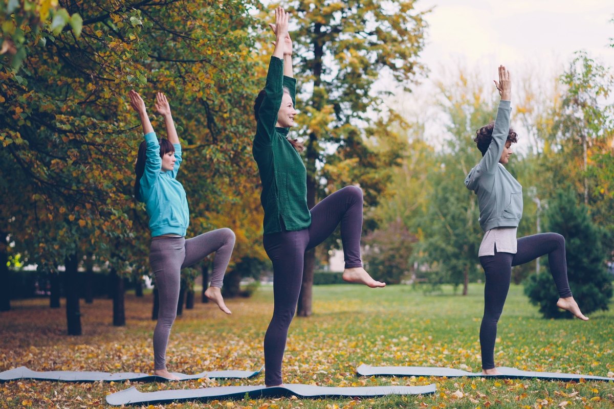 Three women balancing on one leg with their arms raised on yoga mats in a park in autumn