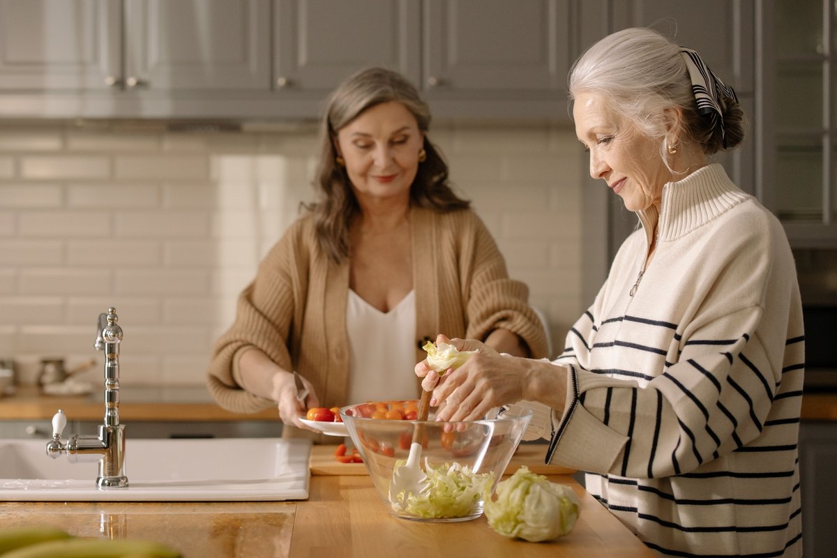 Two older women preparing a salad together at a kitchen island, one slicing tomatoes and one tossing lettuce in a glass bowl