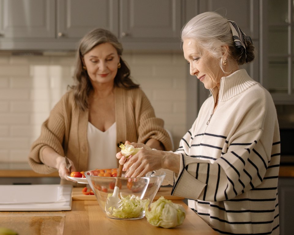 Two older women preparing a salad together at a kitchen island, one slicing tomatoes and one tossing lettuce in a glass bowl